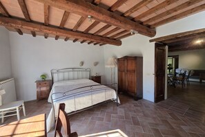A bedroom with a bed, wooden headboard, a nightstand, a lamp, a wardrobe, and a dining area in the background. - Villa Tufi by MMega (Montone)