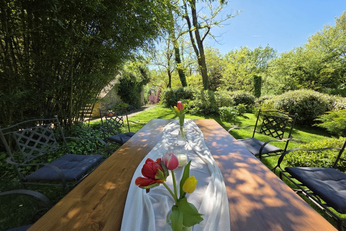 Una mesa de madera con mantel blanco, rodeada de sillas, en un jardín exuberante con árboles y arbustos.