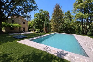Uma piscina retangular com borda de pedra, rodeada por um gramado bem cuidado e árvores.
