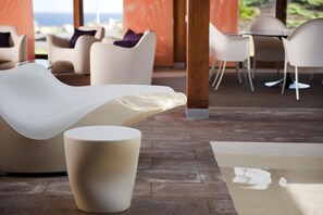 Ein moderner Lounge-Bereich mit einem gebogenen weißen Sofa und einem runden weißen Hocker, mit Blick auf das Meer im Hintergrund.