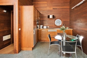 A modern kitchen with wooden cabinetry, a round window, and a dining area with a glass table and metal chairs. - Casas Bioclimáticas Iter El Rio (Granadilla de Abona)