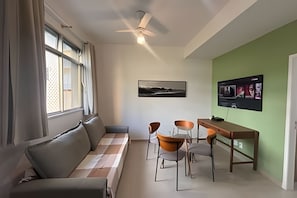 Una sala de estar moderna con un sofá, una mesa de comedor y sillas. Hay un ventilador de techo, una ventana con cortinas y un televisor montado en la pared.