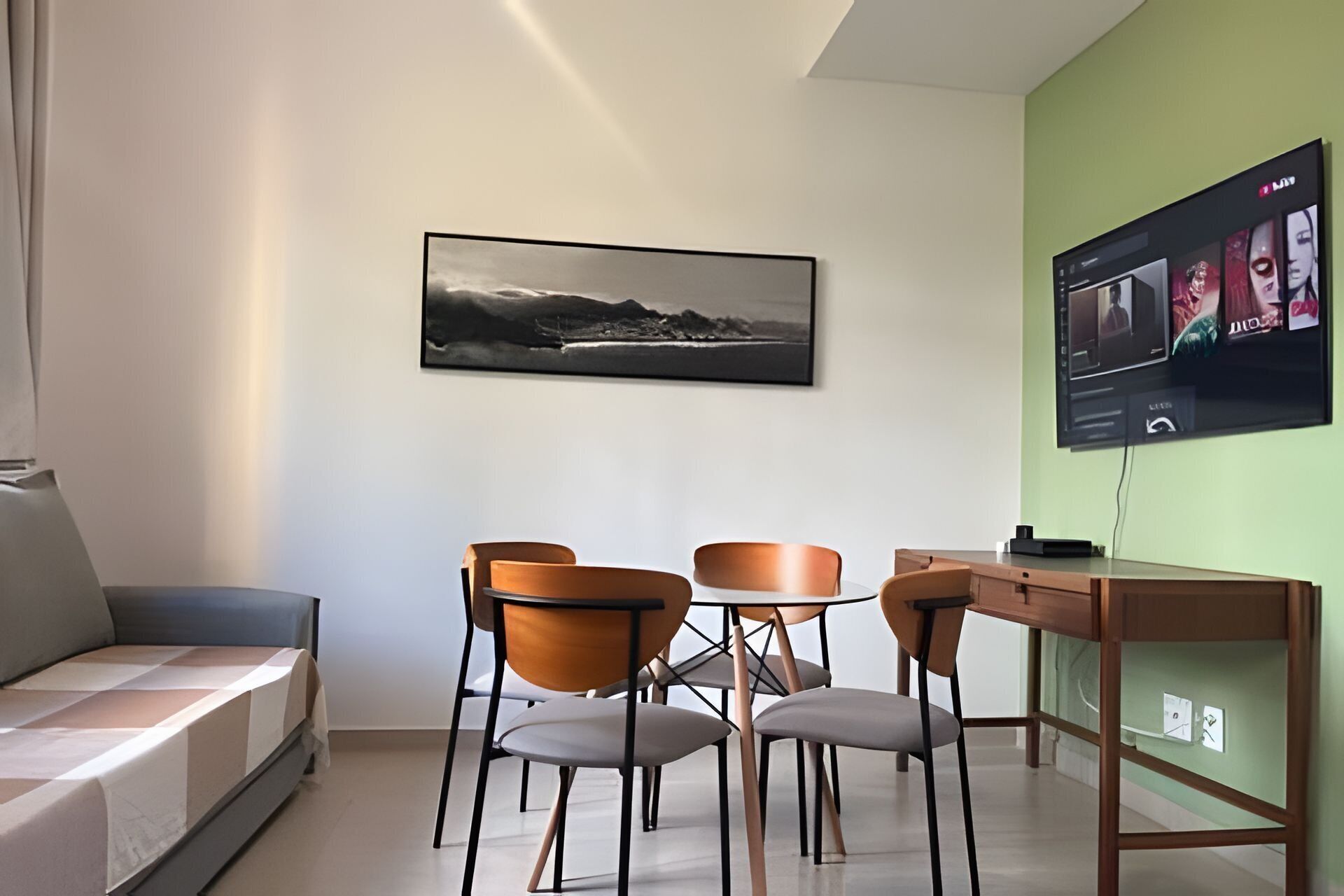 A modern room with a sofa, a dining table with chairs, a flat-screen TV mounted on the wall, and a framed landscape photograph.