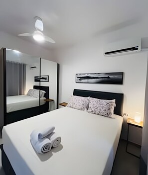 Ein Hotelzimmer mit einem großen Bett, einem Deckenventilator, einer Klimaanlage, einem Nachttisch mit Lampe und einem gerahmten Bild an der Wand.