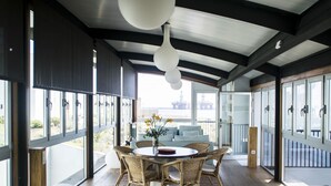 A spacious room with a round table and wicker chairs, large windows with blinds, and a view of an offshore platform. - Casas Bioclimáticas Iter La Religa (Granadilla de Abona)