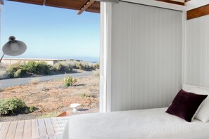 A bedroom with a bed, a window with a view of a road and vegetation, and a lamp on a wooden table. - Casas Bioclimáticas Iter El Escudo (Granadilla de Abona)