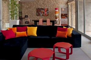 A modern living room with a dark sofa, red and yellow cushions, a red stool, and a stone wall with a painting. - Casas Bioclimáticas Iter El Bernegal (Granadilla de Abona)