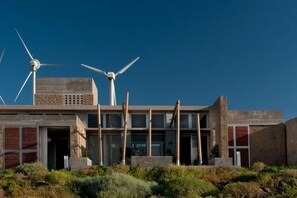 A modern building with multiple wind turbines, a flat roof, and a desert landscape. - Casas Bioclimáticas Iter El Bernegal (Granadilla de Abona)