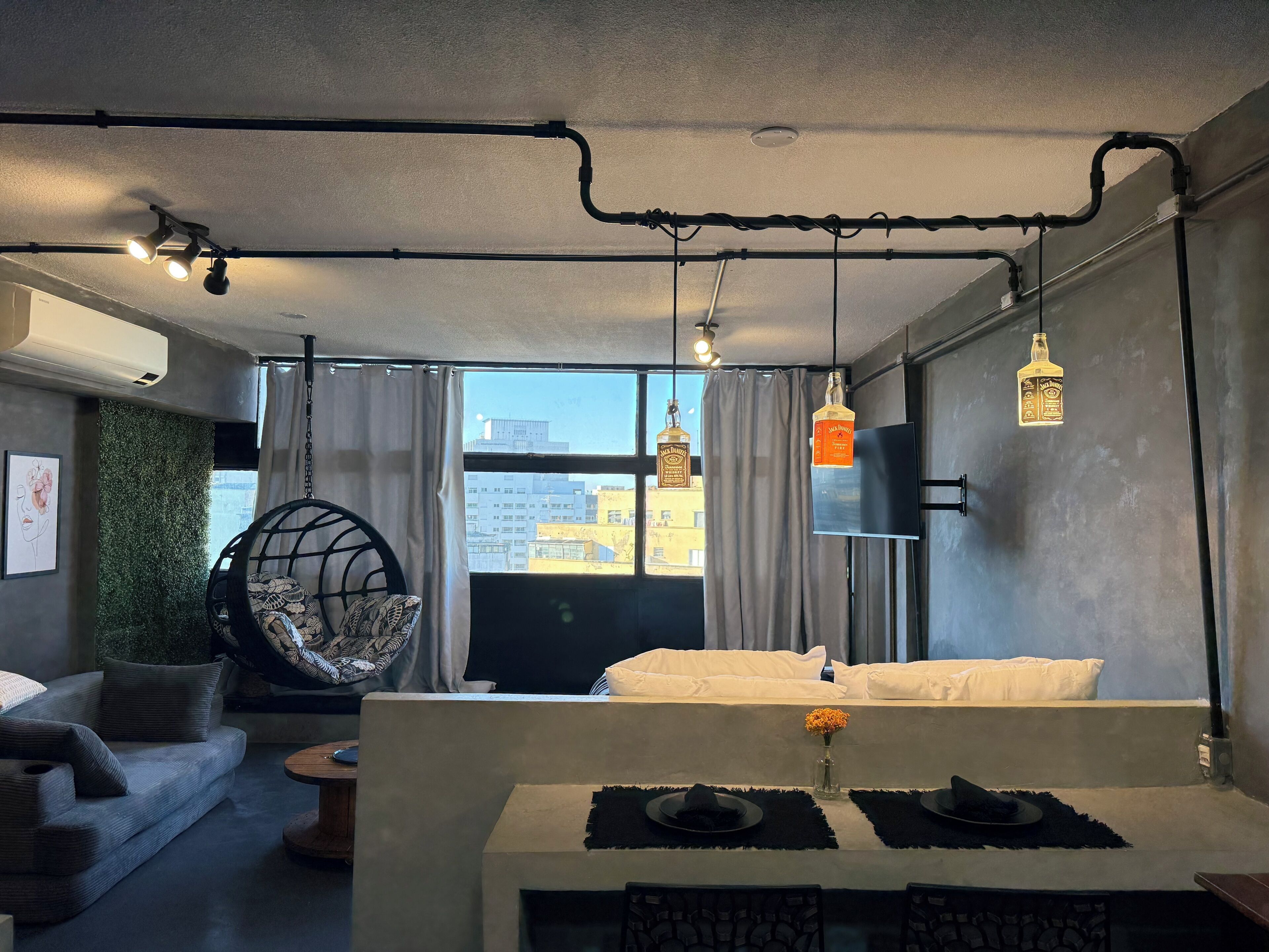 Een moderne hotelkamer met een bed, een hangstoel, een eettafel met een bloemenvaas, en uitzicht op de stad door het raam.