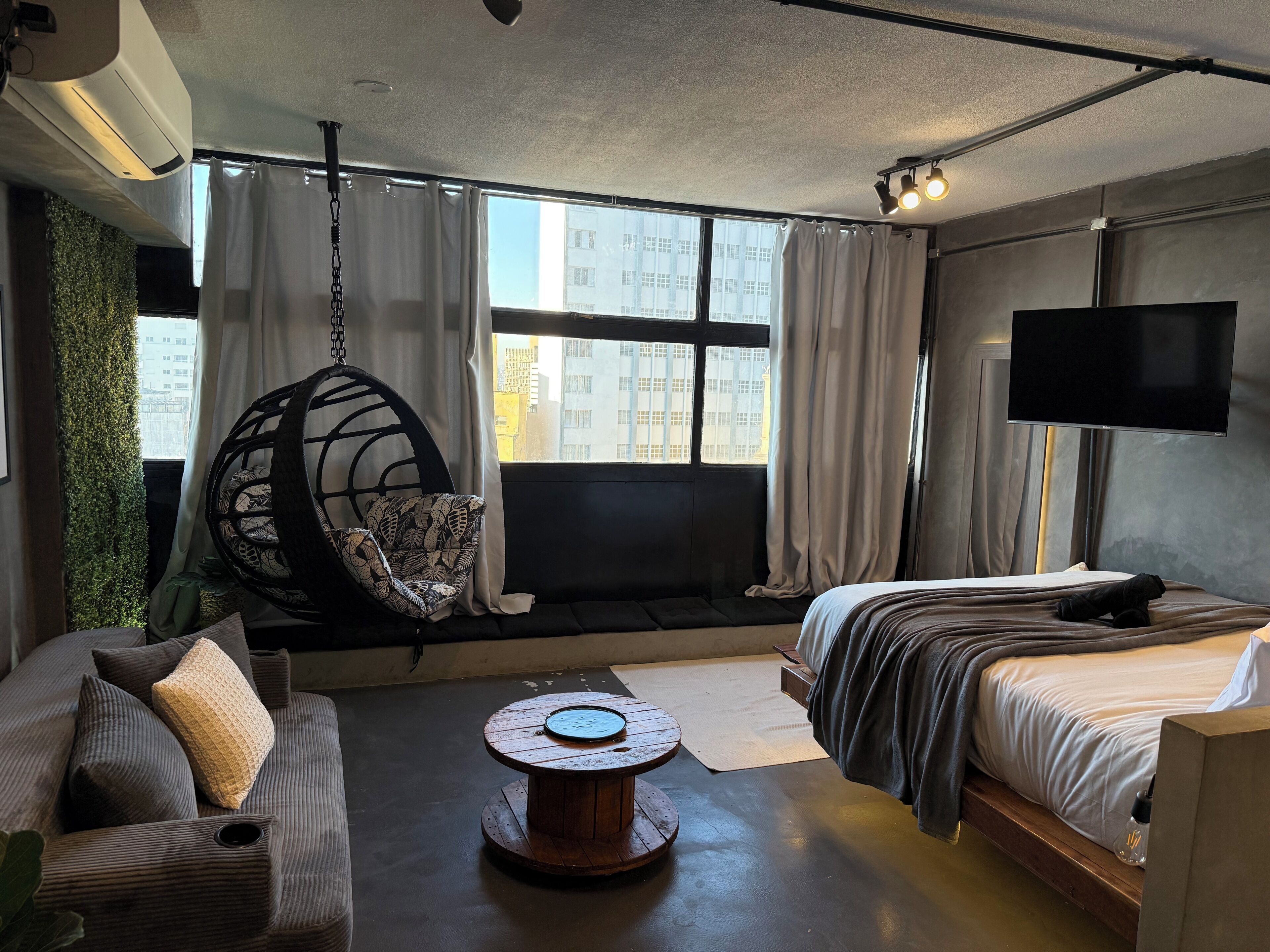 A modern hotel room with a hanging chair, a sofa, a round wooden table, a bed, and a TV.
