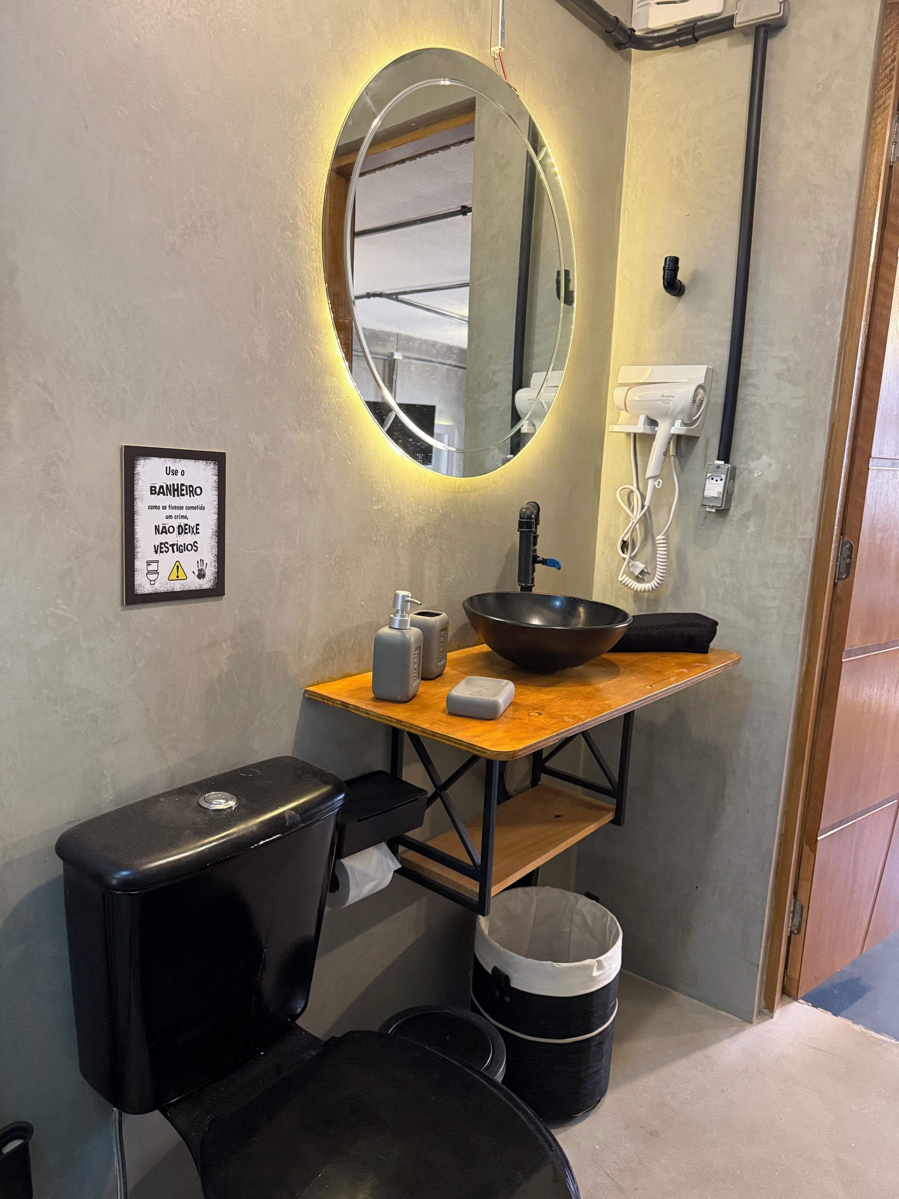 A bathroom with a round mirror, a sink with a black basin, a toilet, and a trash bin.