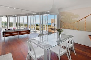 Interior moderno com mesa de jantar de vidro, cadeiras brancas, um banco e vista para o exterior através de grandes janelas.
