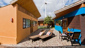 A modern building with a sloped roof, a stone patio with lounge chairs, and a wind turbine in the background. - Casas Bioclimáticas Iter Noche y Dia (Granadilla de Abona)