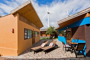 A modern building with a sloped roof, a stone patio with lounge chairs, and a wind turbine in the background. - Casas Bioclimáticas Iter Noche y Dia (Granadilla de Abona)