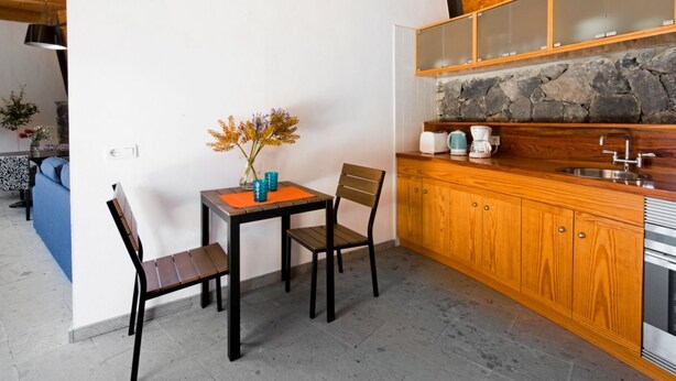 A compact kitchen with a stone backsplash, wooden cabinets, and a dining area with a table and chairs. - Casas Bioclimáticas Iter Noche y Dia (Granadilla de Abona)