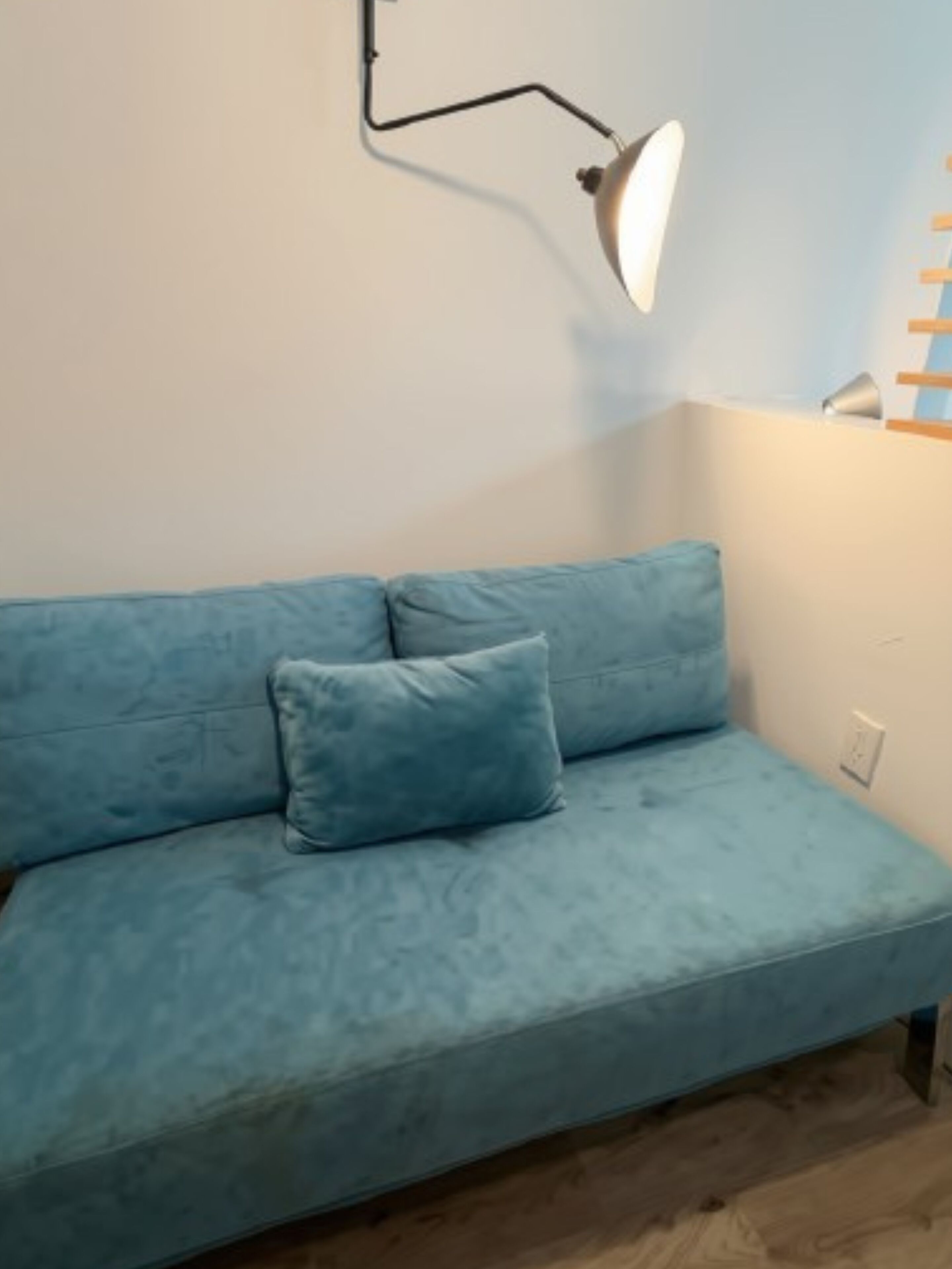 Ein hellblaues Sofa mit einem einzelnen Kissen, eine weiße Wand, eine schwarze Stehlampe und eine Holzleiter, die an der Wand lehnt.