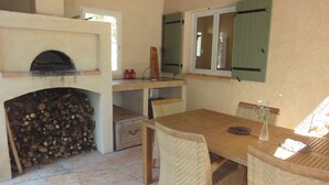 A rustic kitchen with a wood-burning stove, a dining table with wicker chairs, and a window with shutters. - Holiday Home “Waterballet” with Pizza Oven, Wifi, and Air Conditioning (Villecroze)
