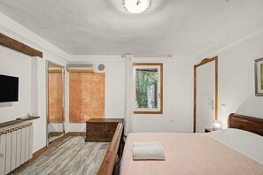 A bedroom with a bed, a wooden headboard, a window with a view of trees, a wall-mounted heater, and a flat-screen TV. - Apartment 'Camera Giuly' with Shared Garden, Wi-Fi and Air Conditioning (Venice)