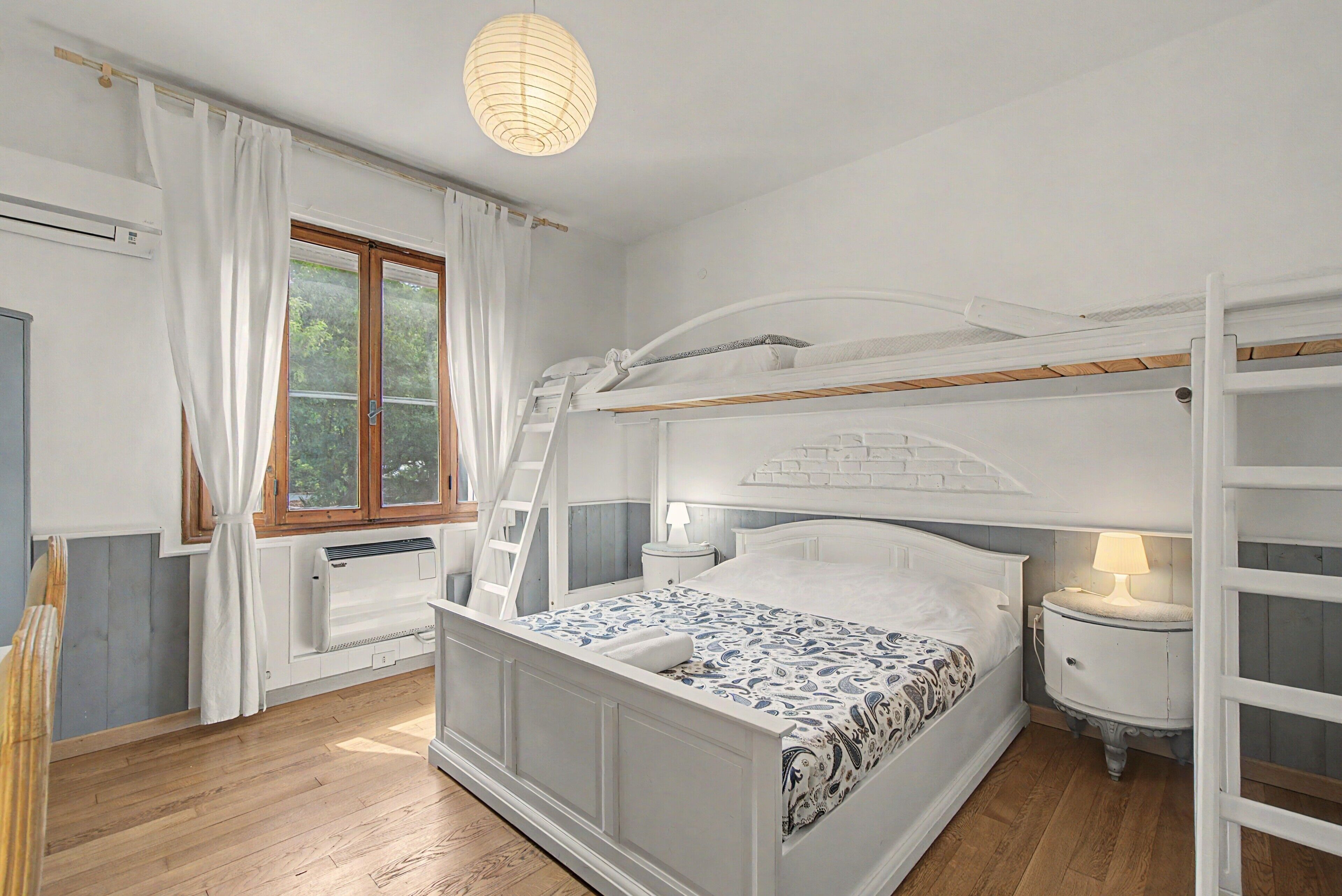 A bedroom with a bunk bed, a bedside table with a lamp, a window with curtains, and an air conditioning unit.