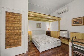 Ein Schlafzimmer mit Holztür, einem Bett mit gemustertem Überwurf, einem Nachttisch, einer Lampe, einer Klimaanlage und einem gerahmten Gemälde an der Wand.