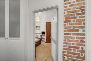 A hallway leading to a bedroom with a brick wall, a bed, a desk, and a television. - 'Camera Rose' Apartment with Shared Garden, Wi-Fi, and Air Conditioning (Venice)