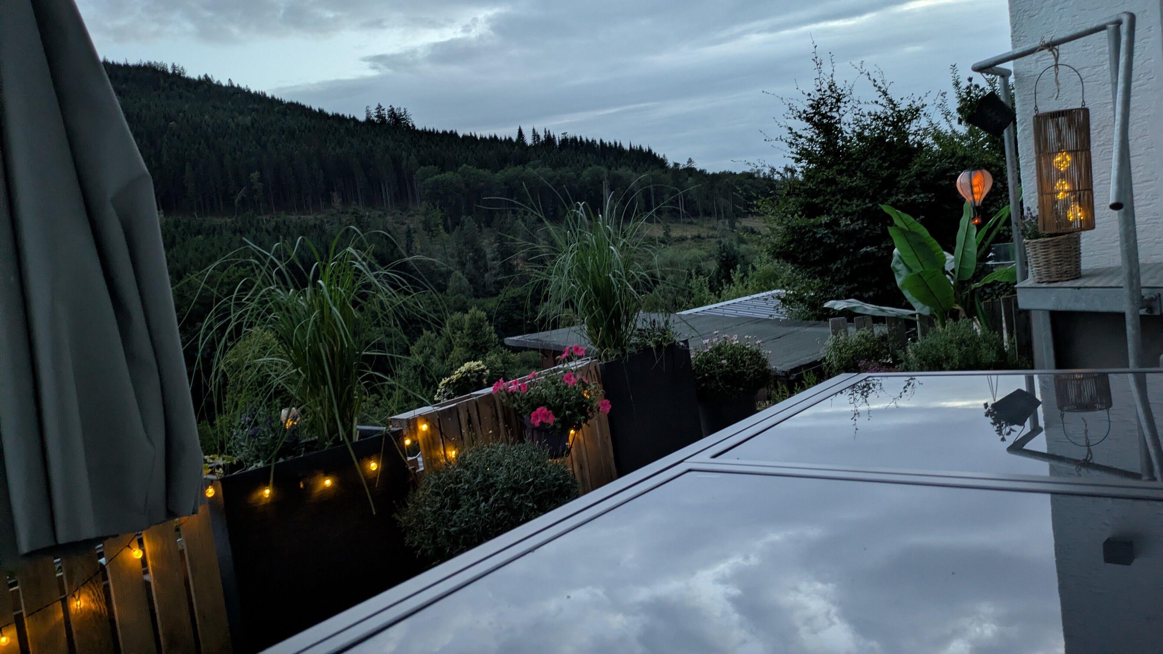 Ein Wohngebiet mit Blick auf einen bewaldeten Hügel, eine Holzterrasse mit Lichterketten und ein Haus mit Satteldach.