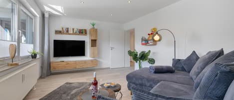 Una sala de estar moderna con un televisor de pantalla plana, una estantería, un sofá y un comedor con mesa y sillas.