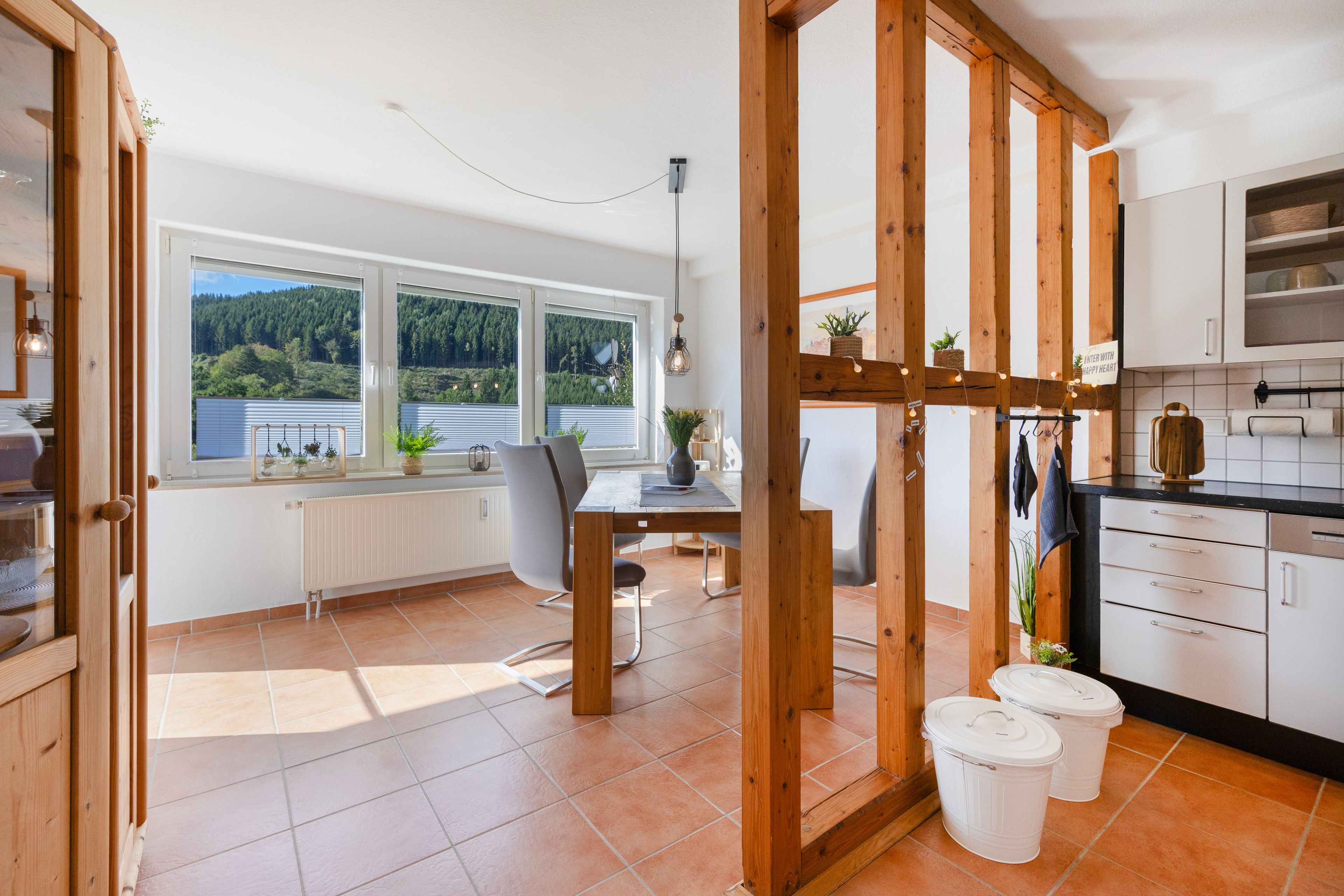 A modern kitchen with wooden beams, a dining area with a view of the forest, and a window with blinds.