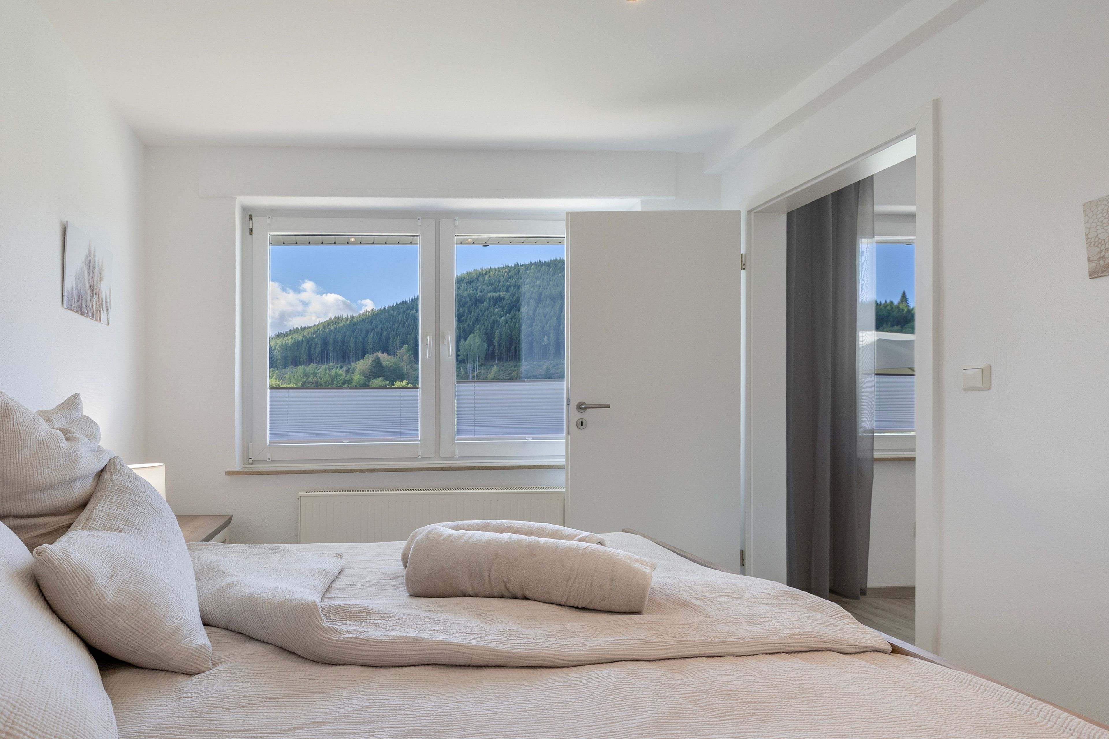 A bedroom with a large bed, a window with a view of a forest and lake, and a door leading to another room.