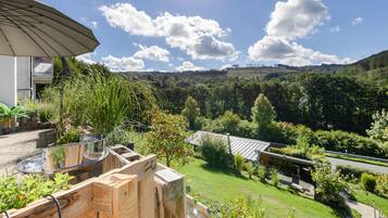 A patio with a glass-top table, wooden deck, and a view of a green landscape with trees and hills.