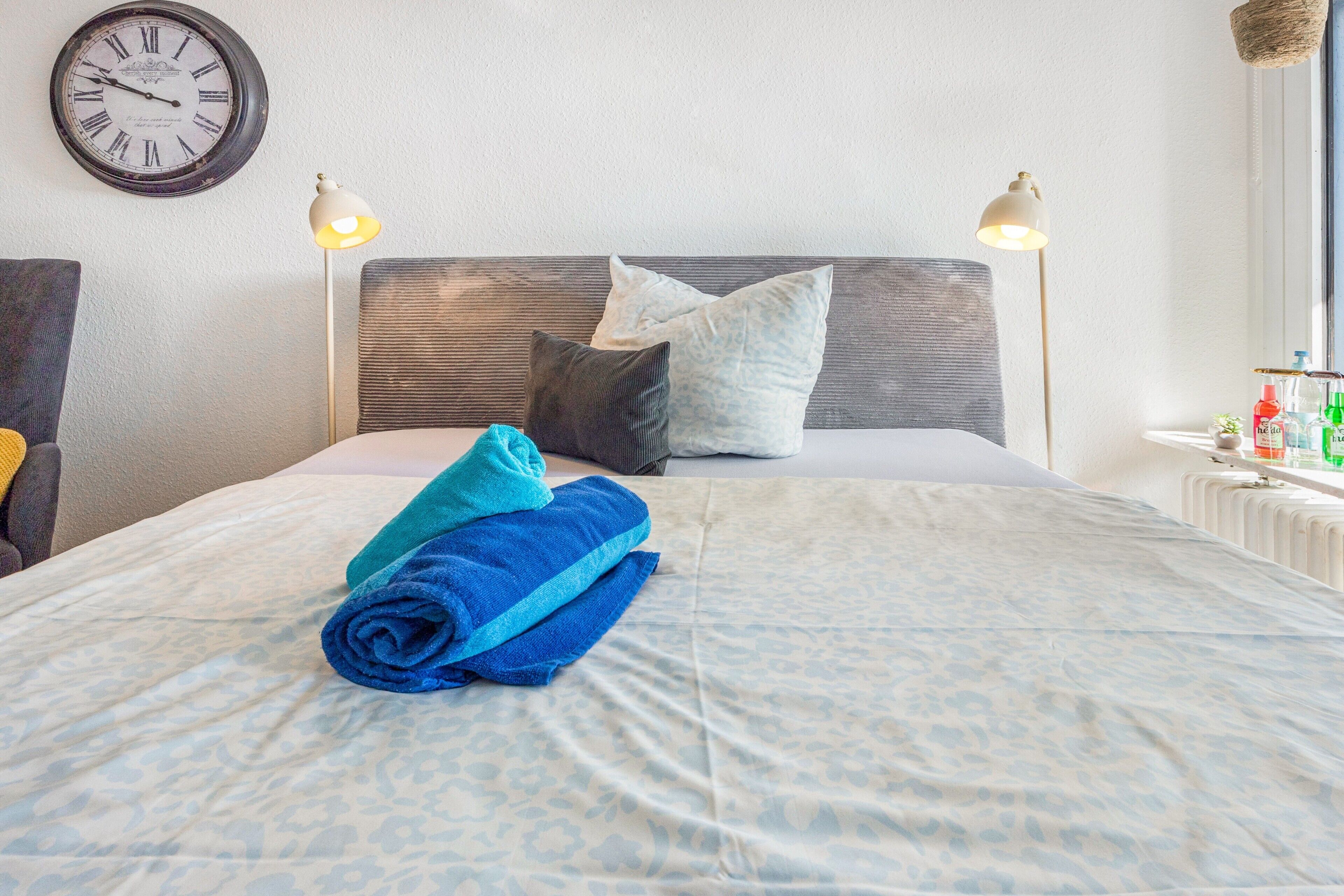 A neatly made bed with a patterned bedspread, two pillows, and rolled towels on a headboard.