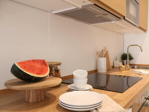 A kitchen with a wooden countertop, a watermelon on a wooden stand, a jar of pasta, and a sink with a faucet.