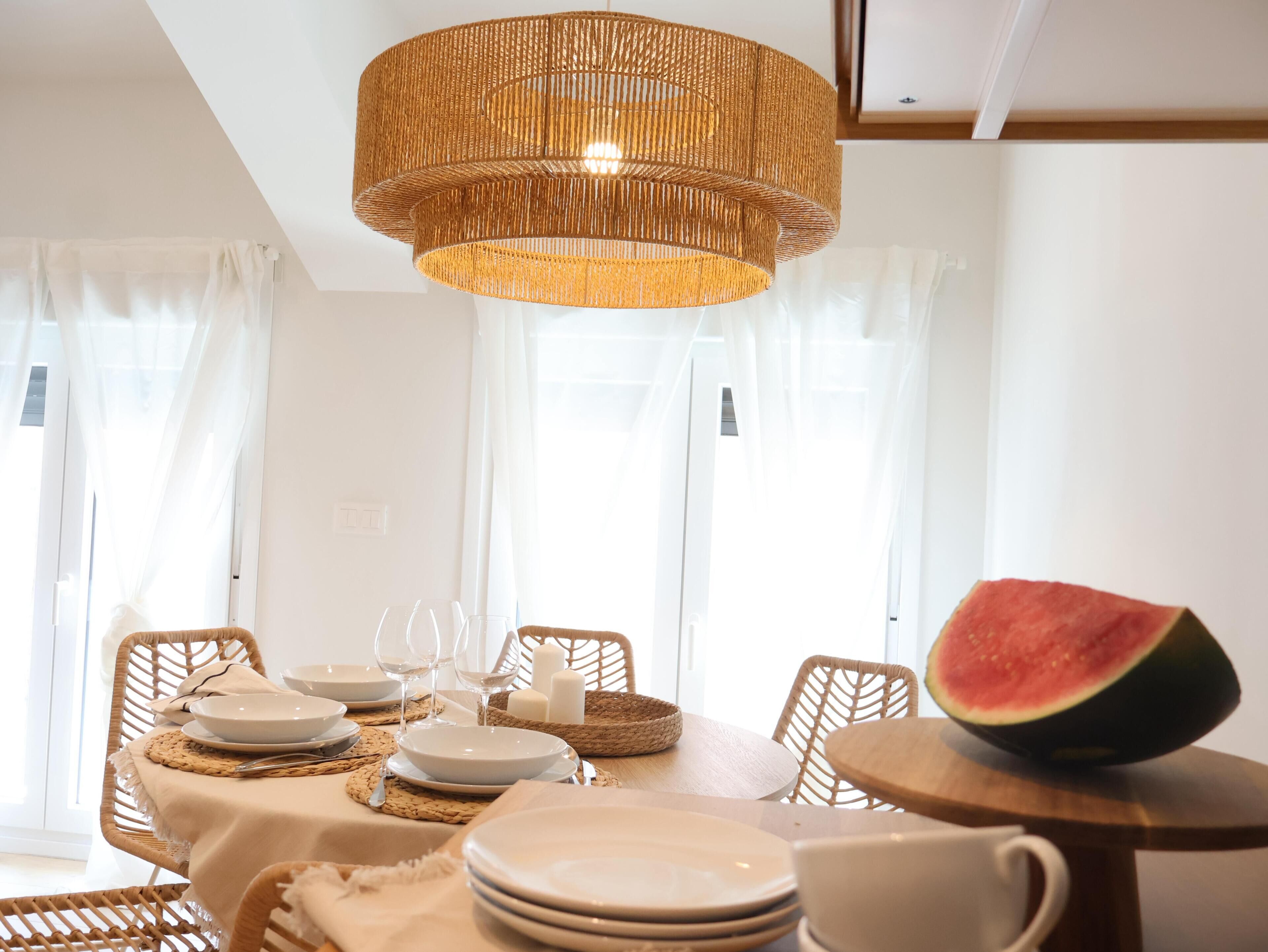 Uma sala de jantar com uma mesa posta para quatro pessoas, com cadeiras de fibra natural, uma luminária pendente trançada e um centro de mesa com frutas.
