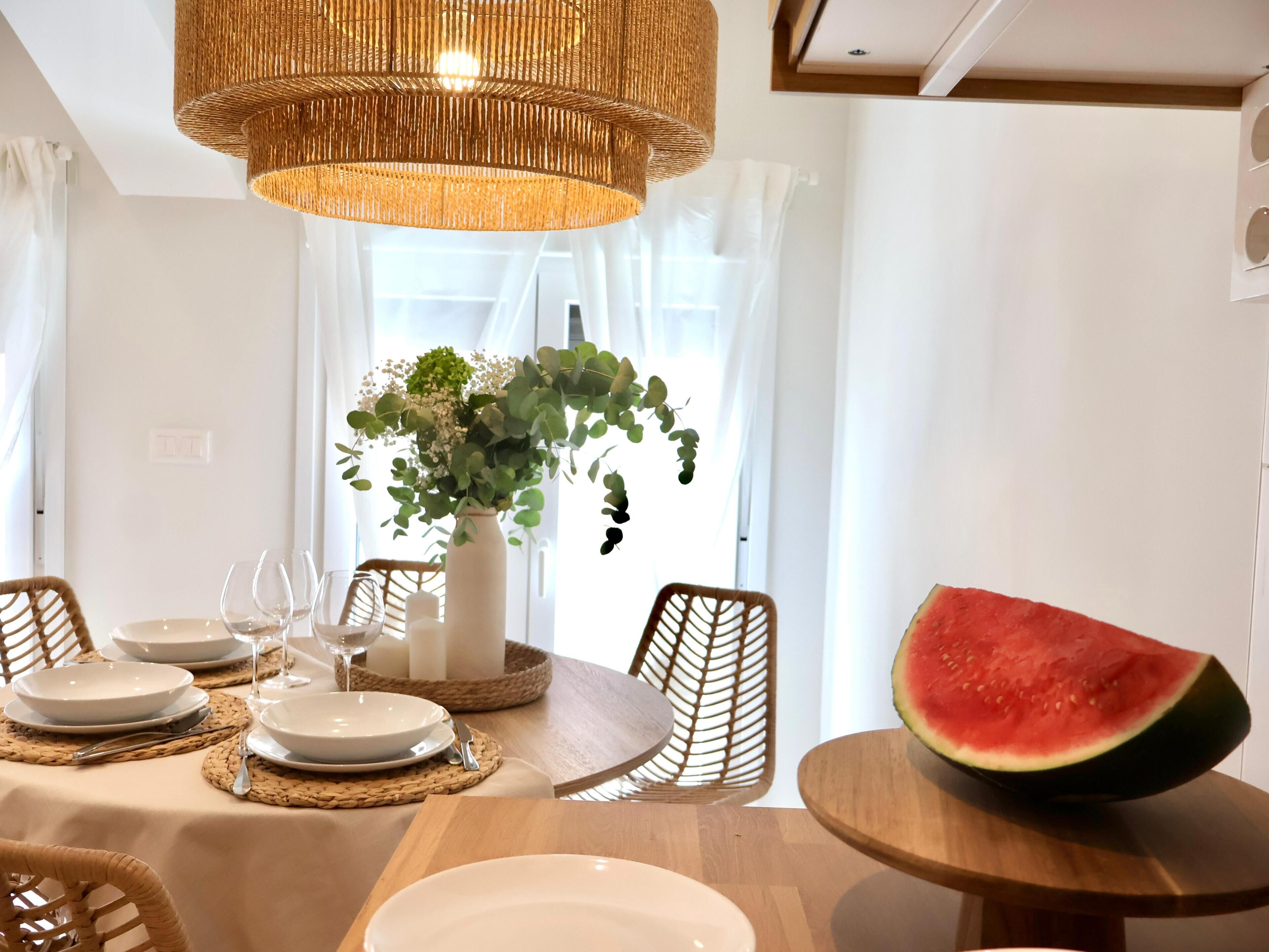 Un coin repas avec une table dressée pour quatre personnes, un vase rempli de fleurs et une grande tranche de pastèque posée sur une table d’appoint.