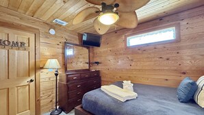 Ein Zimmer mit Holzwandverkleidung, einem Bett, einem Schminktisch, einem Spiegel, einem Deckenventilator und einem Fernseher.