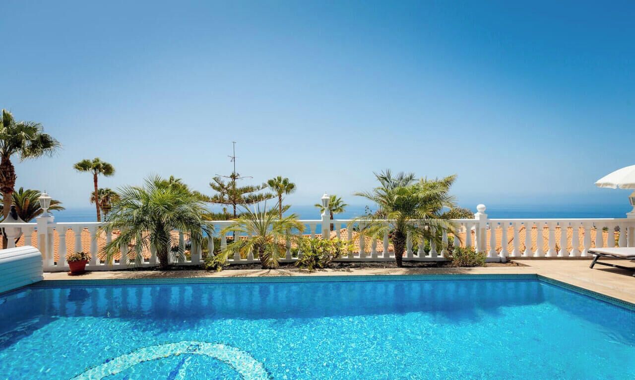A clear blue swimming pool with a white fence, palm trees, and a view of the sea.