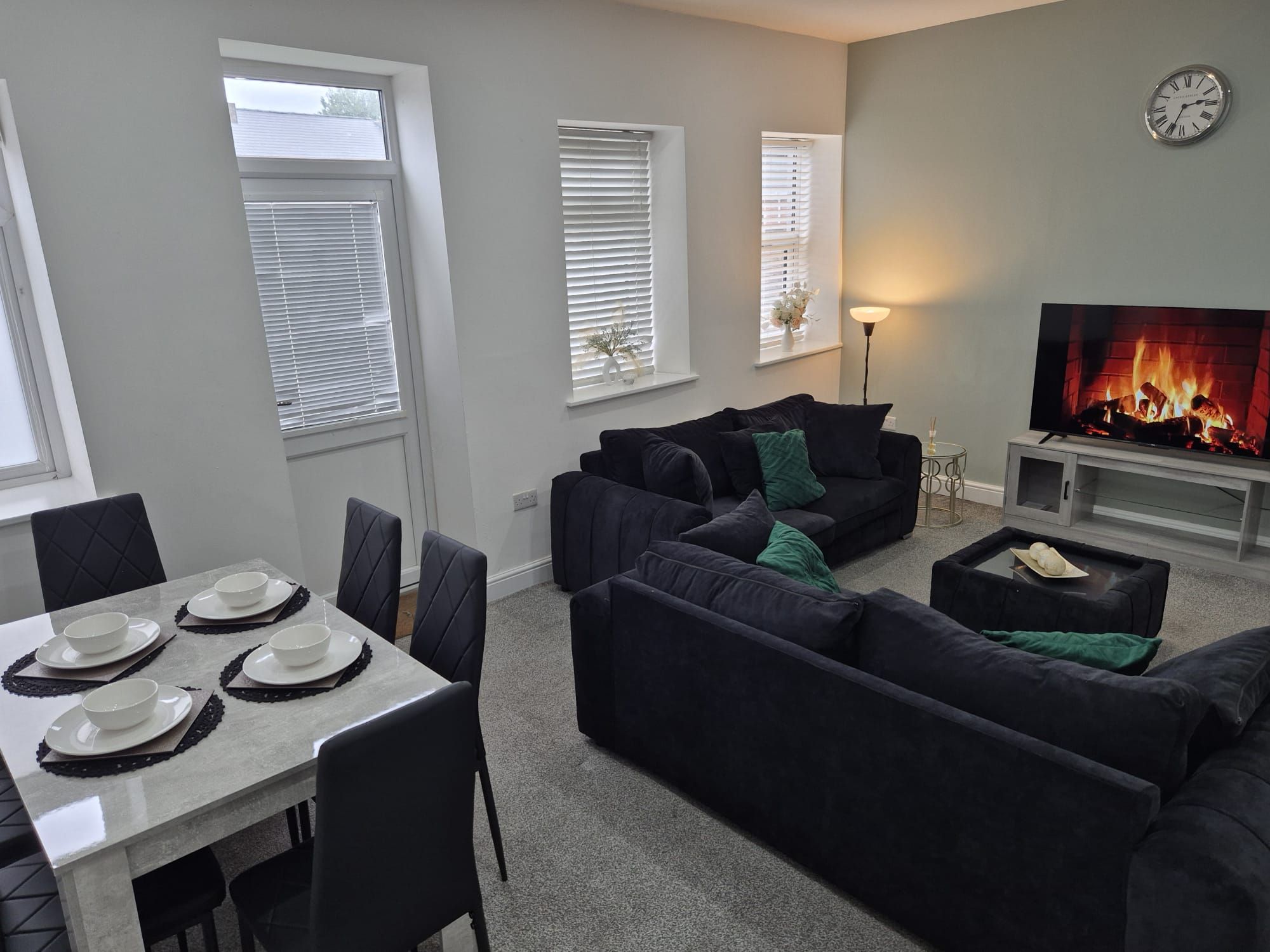 A living room with a dark-colored sofa set, a flat-screen TV, and a fireplace. There is a dining table with plates and cutlery.