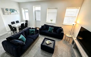 A living room with two dark sofas, a coffee table, a dining table with chairs, and a TV. - 3 Bed House-Sleeps 8-Families-Contractors-NHS-Garden-Parking-Fast WiFi (Willenhall)