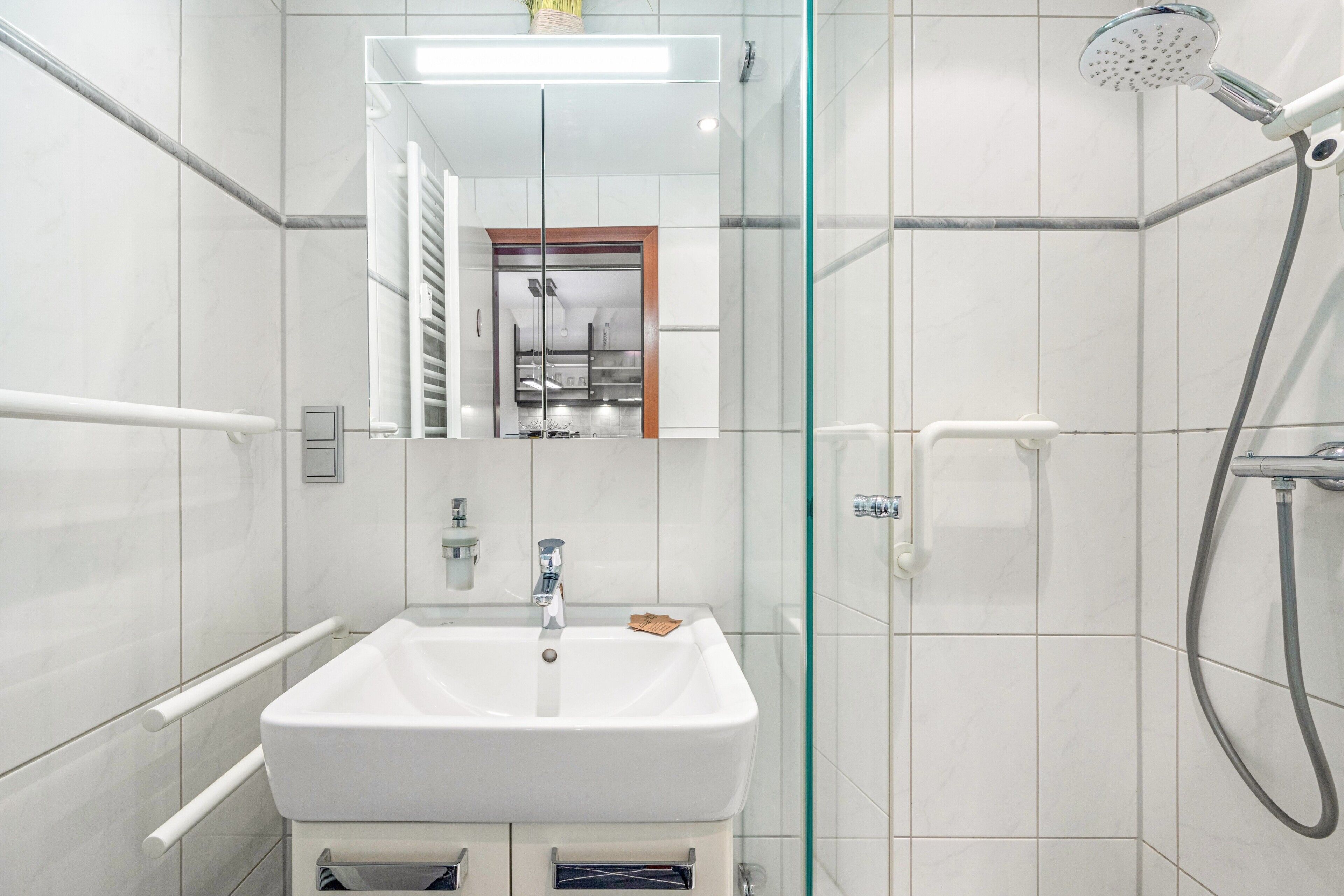 Ein modernes Badezimmer mit einem weißen Waschbecken, einer Duschkabine aus Glas und einem in der Spiegelung erkennbaren Küchenbereich.