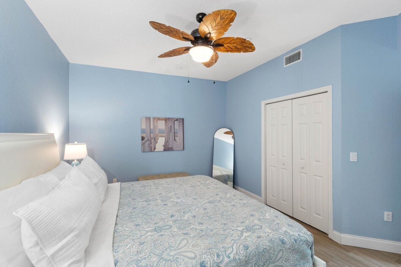 A bedroom with a bed, a ceiling fan, a nightstand, a lamp, a door, and a wall-mounted mirror.