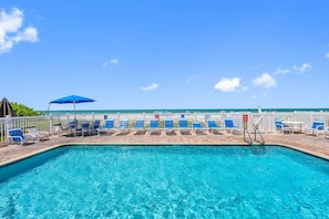 A clear blue pool with lounge chairs and umbrellas, surrounded by a white fence, with a view of the ocean. - Ocean Side Beach Front Penthouse! (Indian Rocks Beach)