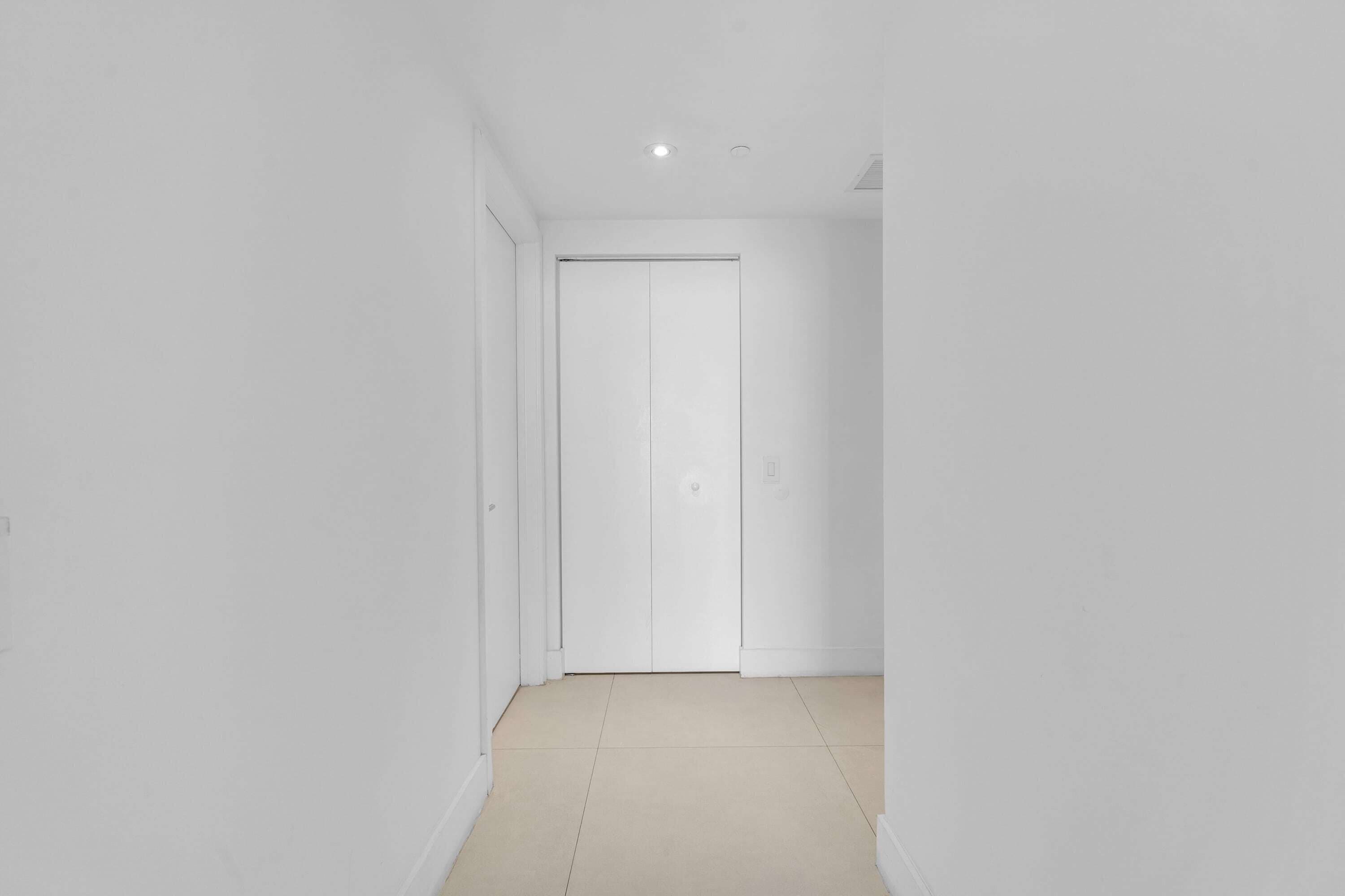 A narrow hallway with a closed white door at the end, white walls, and a tiled floor.