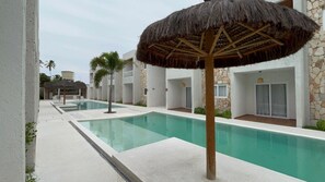 A swimming pool area with a thatched umbrella, stone walls, and a palm tree. - São Miguel dos Milagres, Praia do Marcineiro. New condominium near the town center. (São Miguel dos Milagres)