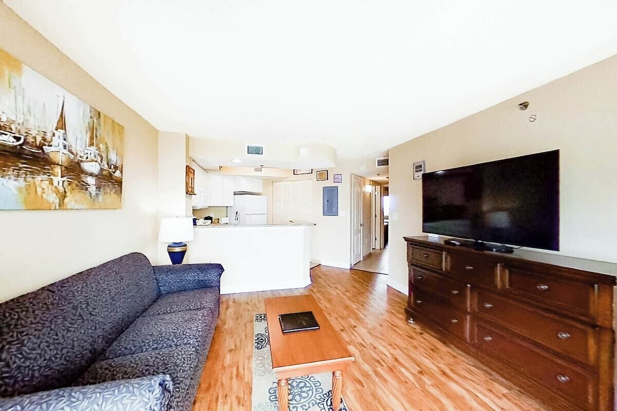 A living room with a sofa, a television, a wooden dresser, and a kitchen in the background.