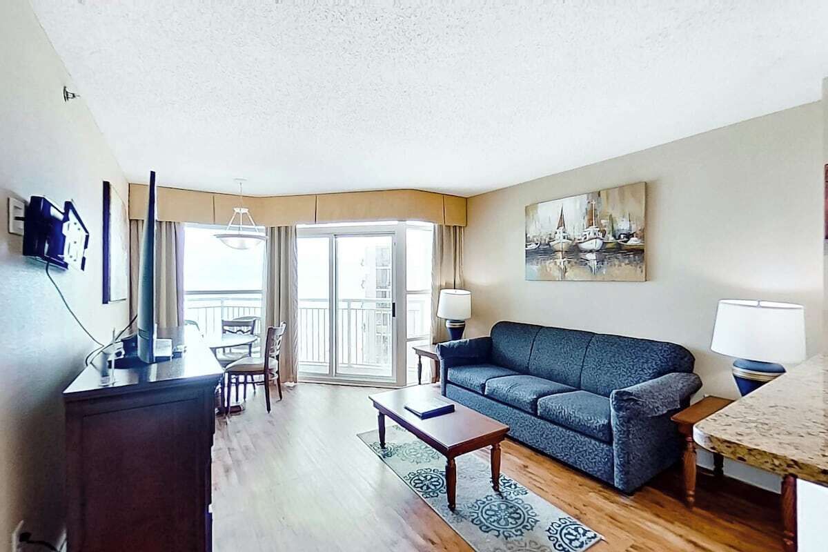 Ein Wohnzimmer mit Sofa, Couchtisch, Fernseher, einem Gemälde und einem Balkon mit Aussicht.