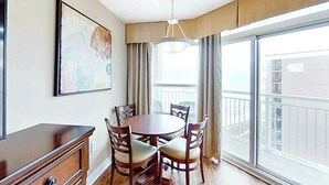 A dining area with a round table and chairs, a pendant light, a painting on the wall, and a view through a large window. - Carolinian Beach Resort 1735 (Myrtle Beach)
