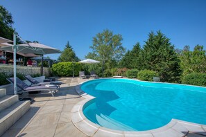 A swimming pool with lounge chairs and umbrellas, surrounded by trees and a stone wall.