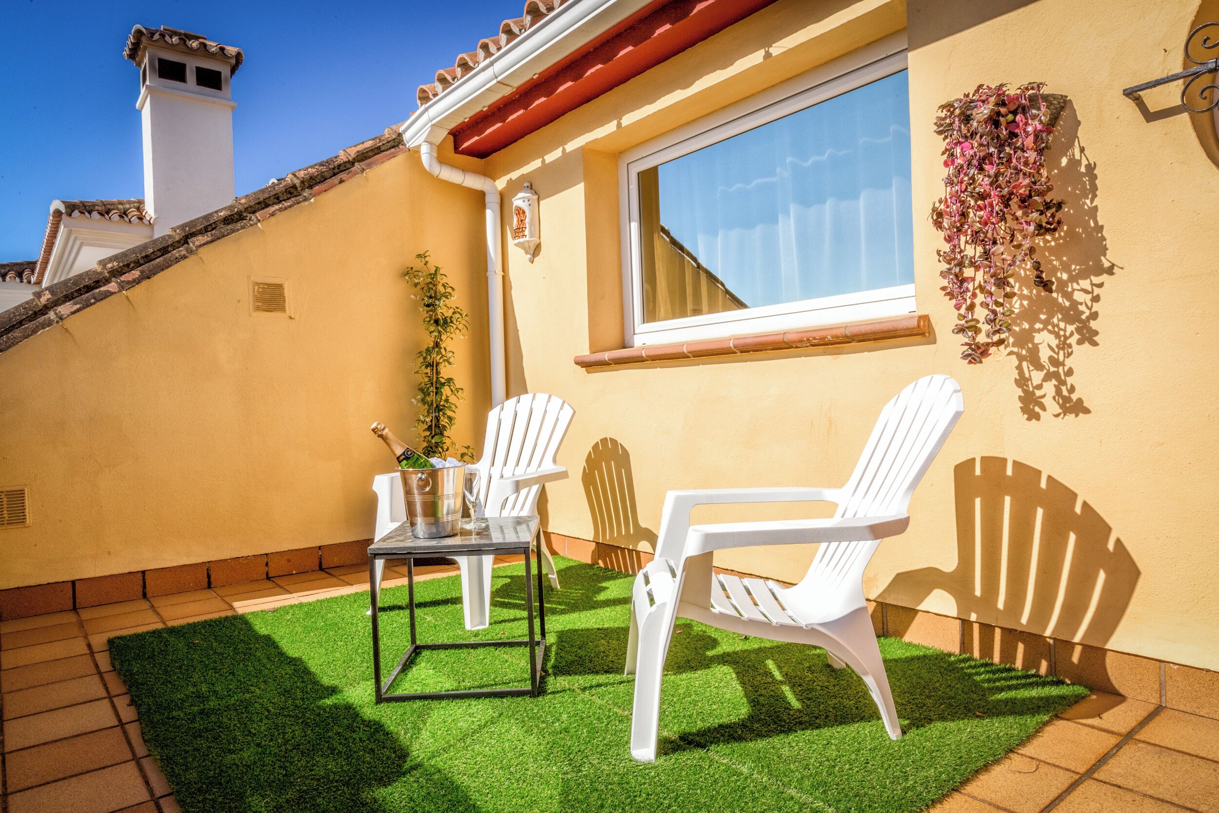 Une terrasse avec deux chaises blanches, une petite table et une bouteille de champagne.