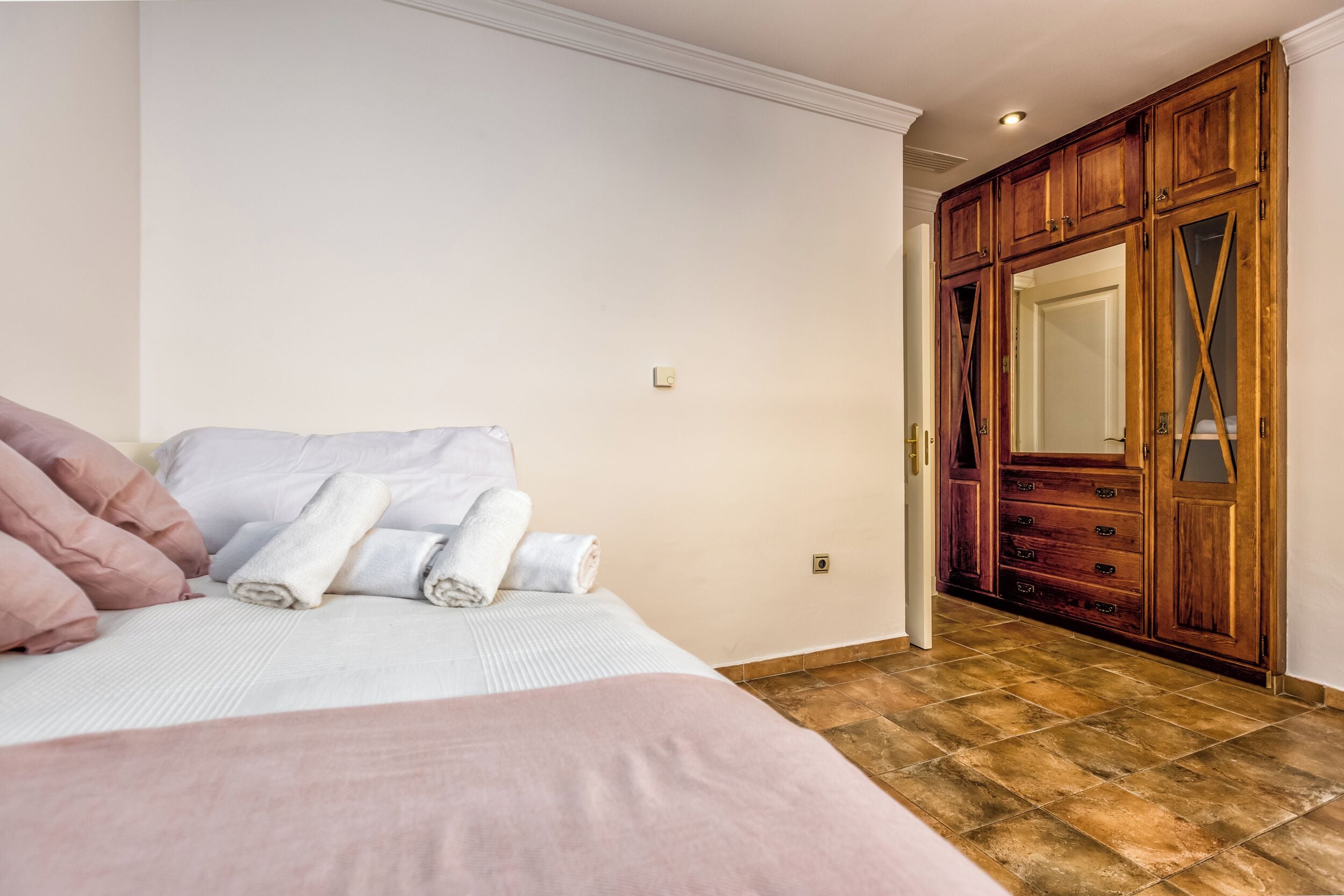 A bedroom with a bed, pillows, and rolled towels. There is a wooden wardrobe with a mirror and a door leading to another room.