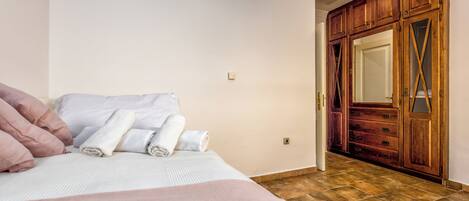 A bedroom with a bed, pillows, and rolled towels. There is a wooden wardrobe with a mirror and a door leading to another room.